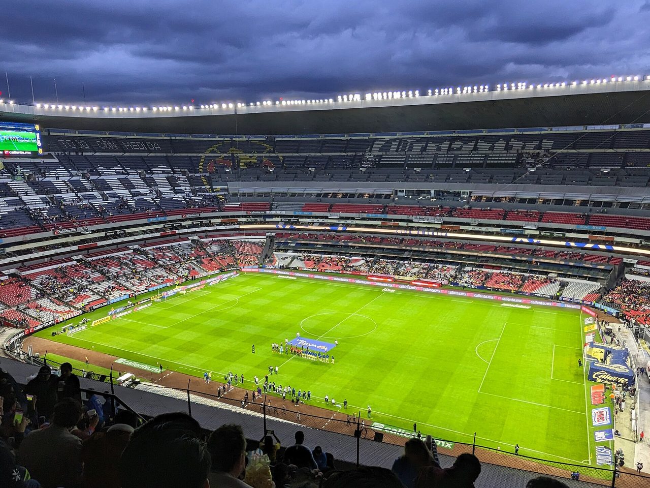 Interior del Estadio Azteca al anochecer, con el césped verde brillante y los equipos formados en el círculo central